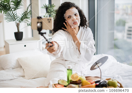 Young female influencer in paper sheet mask holding smartphone Young female influencer in paper sheet mask holding smartphone 109896470