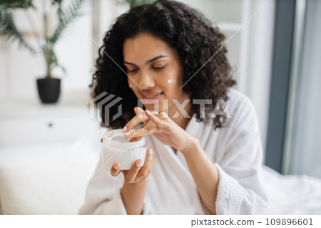 Multiracial woman holding jar with cream on hand and applying on skin 109896601
