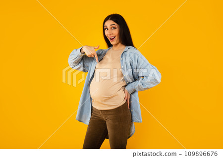 Happy pregnancy. Excited expectant woman pointing at her big belly and smiling at camera, yellow background Happy pregnancy. Excited expectant woman pointing at her big belly and smiling at camera, yellow background 109896676