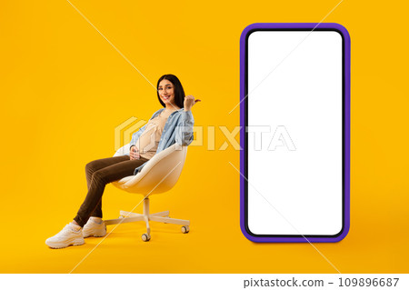 Mobile offer. Positive expectant woman pointing back at big smartphone with blank screen, sitting on chair, mockup Mobile offer. Positive expectant woman pointing back at big smartphone with blank screen, sitting on chair, mockup 109896687