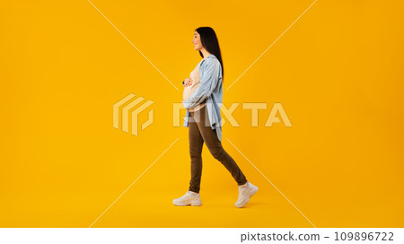 Pregnancy concept. Pregnant lady walking over yellow studio background, full length shot of expectant woman, side view 109896722