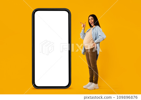 Happy pregnant woman pointing at big smartphone with white screen, standing on yellow studio background, mockup 109896876