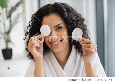 Joyful young african american female in bathrobe covering eye with cotton pad 109897061