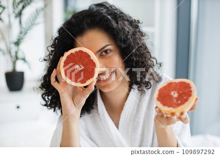 Attractive calm female in white bathrobe covering one eye with grapefruit half Attractive calm female in white bathrobe covering one eye with grapefruit half 109897292