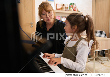 Little girl sitting at piano near her teacher pianist musician, learning music during individual music lesson at home 109897512