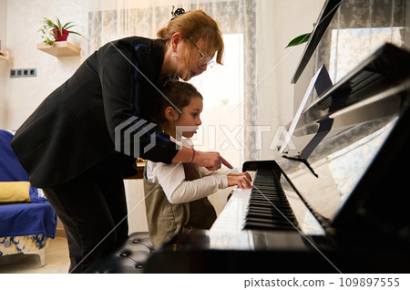 Mature woman pianist teacher giving piano lesson to a little school girl, teaching the position of fingers on piano keys 109897555