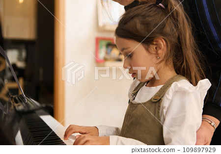 Adorable Caucasian little child girl playing piano under the guidance of her teacher. 109897929