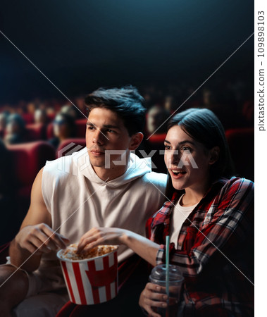 Young man and woman, couple, friends visiting cinema, watching movie with interest and excitement, eating popcorn 109898103