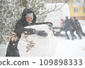a girl is playing with snow on the street, making a snowman, on background a car is stuck in snow and men are pushing, it is snowing, blizzard and frost on a winter day 109898333