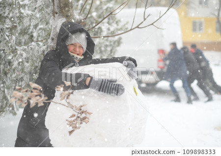 a girl is playing with snow on the street, making a snowman, on background a car is stuck in snow and men are pushing, it is snowing, blizzard and frost on a winter day a girl is playing with snow on the street, making a snowman, on background a car is stuck in snow and men are pushing, it is snowing, blizzard and frost on a winter day 109898333