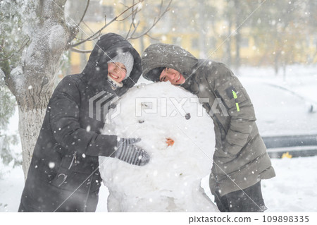a girl and a boy is playing with snow on the street, making snowman, it is snowing, a blizzard and frost on a winter day 109898335