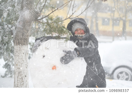 a girl is playing with snow on the street, making snowman, it is snowing, a blizzard and frost on a winter day 109898337
