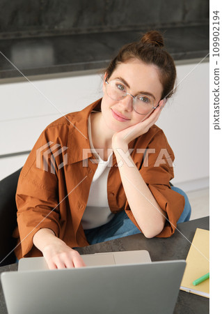 Vertical shot of beautiful woman working from home, student doing homework in kitchen, using laptop, looking at camera and smiling Vertical shot of beautiful woman working from home, student doing homework in kitchen, using laptop, looking at camera and smiling 109902194