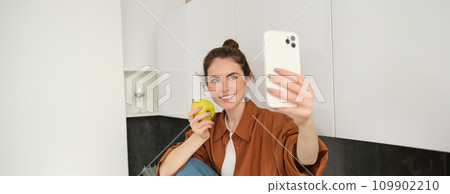 Portrait of happy, smiling young woman records herself, takes selfie while eating an apple in the kitchen, using smartphone app, makes photos with mobile phone Portrait of happy, smiling young woman records herself, takes selfie while eating an apple in the kitchen, using smartphone app, makes photos with mobile phone 109902210