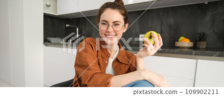 Portrait of smiling, modern woman in glasses, sitting at home and eating green apple, having a lunch break, biting fruit Portrait of smiling, modern woman in glasses, sitting at home and eating green apple, having a lunch break, biting fruit 109902211