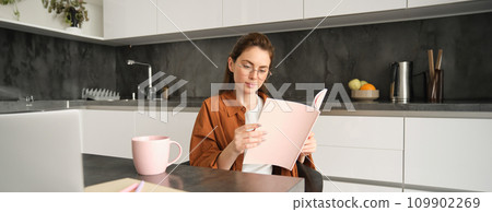 Portrait of woman working from home, sitting in kitchen with documents, opens folder, drinks tea Portrait of woman working from home, sitting in kitchen with documents, opens folder, drinks tea 109902269