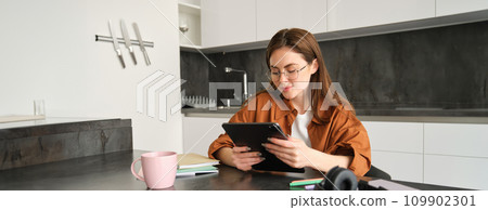 Portrait of beautiful young woman in glasses, reading on digital tablet, watching video, studying at home remotely, sitting in kitchen with headphones, papers and notebooks Portrait of beautiful young woman in glasses, reading on digital tablet, watching video, studying at home remotely, sitting in kitchen with headphones, papers and notebooks 109902301