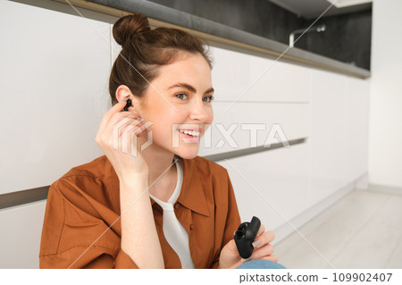 Image of modern brunette girl, sits on kitchen floor, listens to music in wireless headphones, holding earphones and case in hands, looking aside 109902407