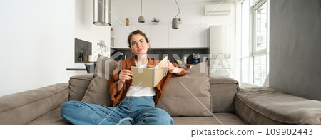 Portrait of young smiling beautiful woman, sitting on sofa in her living room, holding study material, doing homework, reading notes in notebook Portrait of young smiling beautiful woman, sitting on sofa in her living room, holding study material, doing homework, reading notes in notebook 109902443