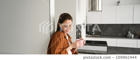 Portrait of cosy, beautiful and relaxed young woman at home, standing and leaning on wall with cup of coffee, drinking tea with thoughtful face expression Portrait of cosy, beautiful and relaxed young woman at home, standing and leaning on wall with cup of coffee, drinking tea with thoughtful face expression 109902444