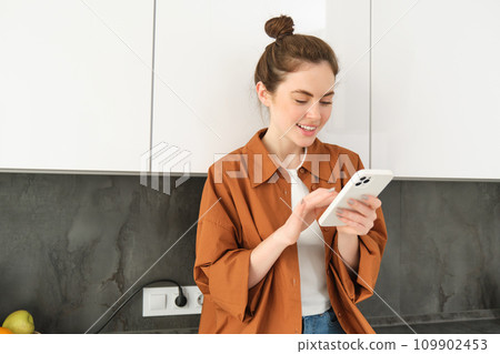 Image of beautiful young female model, posing with smartphone in the kitchen, looking at mobile screen and smiling while reading a message on phone 109902453