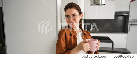 Portrait of beautiful, smiling female model, woman in glasses with cup of coffee, leans on wall at home, drinks tea Portrait of beautiful, smiling female model, woman in glasses with cup of coffee, leans on wall at home, drinks tea 109902459