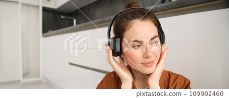 Image of modern woman, beautiful girl enjoys listening to music, has black wireless headphones on, sits on floor and rests 109902460