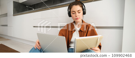 Portrait of young woman, modern student sits at home on floor and doing homework on laptop, listens music in headphones while making assignment or project for college 109902468