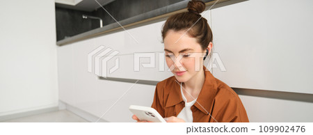 Portrait of beautiful smiling woman sitting on kitchen floor, scrolling social media on mobile phone, listening music in wireless headphones 109902476
