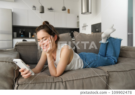 Portrait of cute young woman with smartphone, lying on couch and resting at home, scrolling social media feed, online shopping, reading something on mobile phone 109902478
