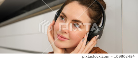 Close up portrait of beautiful brunette woman, listens to music in wireless headphones, has new earphones on her head, smiling and looking happy Close up portrait of beautiful brunette woman, listens to music in wireless headphones, has new earphones on her head, smiling and looking happy 109902484
