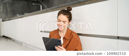 Portrait of young beautiful woman sitting on kitchen floor with digital tablet, browsing news feed, social media app on gadget, smiling and looking happy Portrait of young beautiful woman sitting on kitchen floor with digital tablet, browsing news feed, social media app on gadget, smiling and looking happy 109902510