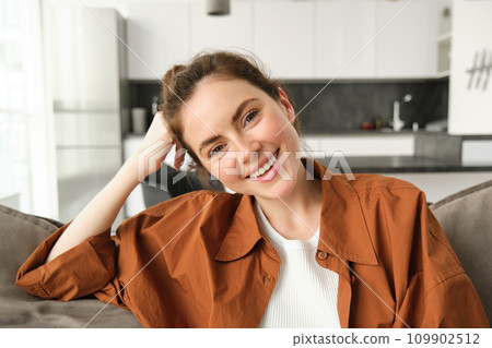 Close up shot of smiling beautiful young woman sits on sofa, resting on couch and looking happy at camera, watching television at home Close up shot of smiling beautiful young woman sits on sofa, resting on couch and looking happy at camera, watching television at home 109902512