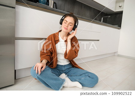 Technology concept. Young smiling brunette woman in wireless headphones, listens to music, sits on kitchen floor Technology concept. Young smiling brunette woman in wireless headphones, listens to music, sits on kitchen floor 109902522