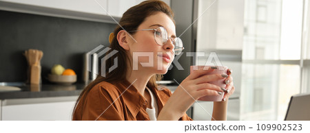 Portrait of young woman enjoys quite day at home with hot cup of tea. Female model sits in kitchen with mug of aromatic coffee, wearing glasses, looking aside Portrait of young woman enjoys quite day at home with hot cup of tea. Female model sits in kitchen with mug of aromatic coffee, wearing glasses, looking aside 109902523