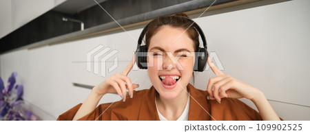 Cute young woman, makes funny faces, listens to music, wears wireless headphones, having fun at home 109902525