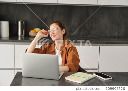 Portrait of young woman, business owner in glasses, sitting with laptop in kitchen, working from home. Student studying, doing homework on computer Portrait of young woman, business owner in glasses, sitting with laptop in kitchen, working from home. Student studying, doing homework on computer 109902528