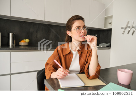 Young creative woman, writer making notes, holding pen, looking aside with thinking face, sitting in kitchen at home, doing homework 109902531