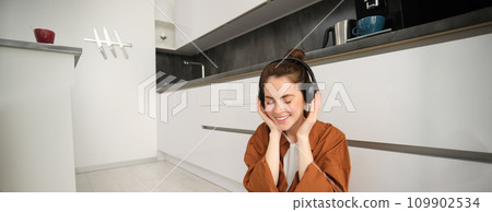 Portrait of beautiful female model on kitchen floor at home, listens music in wireless headphones, smiling and looking happy Portrait of beautiful female model on kitchen floor at home, listens music in wireless headphones, smiling and looking happy 109902534