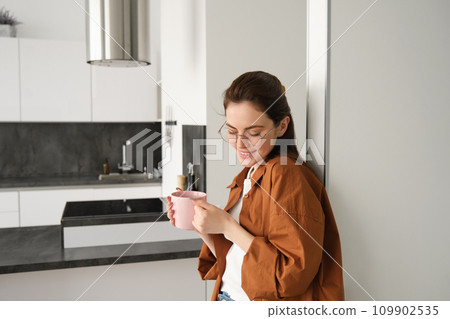 Portrait of cosy, beautiful and relaxed young woman at home, standing and leaning on wall with cup of coffee, drinking tea with thoughtful face expression Portrait of cosy, beautiful and relaxed young woman at home, standing and leaning on wall with cup of coffee, drinking tea with thoughtful face expression 109902535