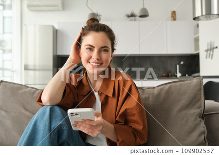 Beautiful smiling female model sitting on couch with smartphone, looking happy at camera, scrolling social media, messaging someone, doing online shopping Beautiful smiling female model sitting on couch with smartphone, looking happy at camera, scrolling social media, messaging someone, doing online shopping 109902537