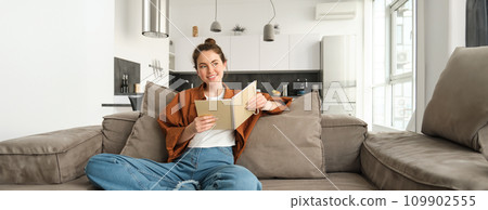 Young woman sits at home on sofa, reading her notes, holding notebook, studying for exam, student revising in her living room 109902555