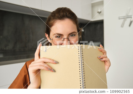 Close up portrait of cute brunette woman in glasses, sitting at home, smiling, hiding face behind yellow notebook 109902565