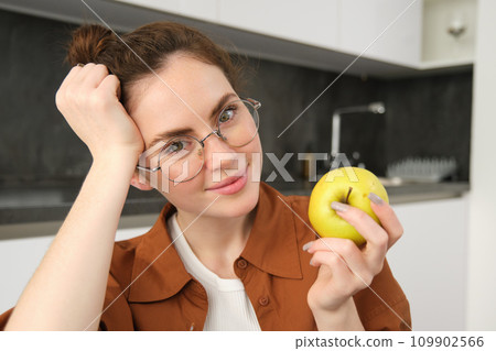 Close-up portrait of brunette woman at home, wearing glasses, eating apple in the kitchen and smiling, biting fruit 109902566