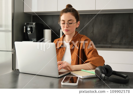 Work and life balance concept. Young woman in glasses, working from home, typing on laptop, student doing homework on computer, sitting in kitchen, freelancing Work and life balance concept. Young woman in glasses, working from home, typing on laptop, student doing homework on computer, sitting in kitchen, freelancing 109902578