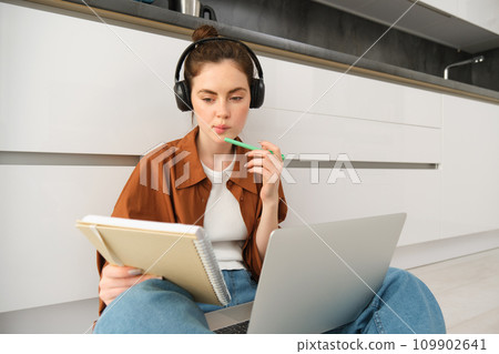 Serious-looking girl student studying at home, sits with laptop and notebook on floor, doing homework project on computer, has online course lesson, wears headphones 109902641