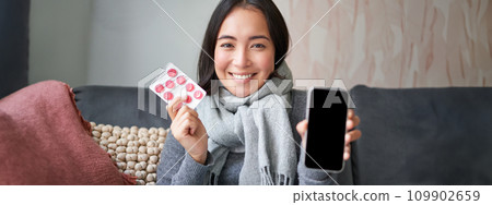 Happy asian woman catching cold, showing smartphone screen and pills, recommending online doctor app, GP or medical clinic on phone, holding drugs, staying at home with flu 109902659
