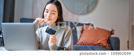 Happy smiling girl with credit card, paying her bills online on computer, doing shopping on her laptop, sitting at home 109902699