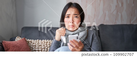 Asian girl with influenza, sits at home, cries and looks upset, holds smartphone, feels unwell and gloomy, wearing warm clothes Asian girl with influenza, sits at home, cries and looks upset, holds smartphone, feels unwell and gloomy, wearing warm clothes 109902707