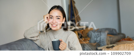 Smiling asian woman sitting on sofa with her mug, drinking coffee at home and relaxing after work, looking calm and cozy 109902709